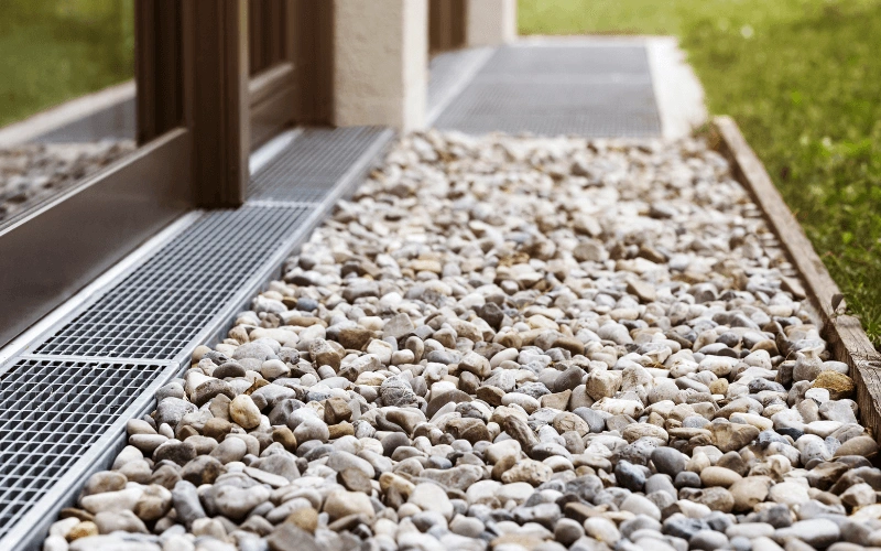 800x450 image of a bed of small rocks and silver drain just outside of a home indicating a french drain for the home underground drainage system