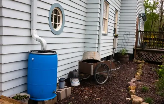 blue rain water collection barrel sitting at the side of a grayish colored home under a white gutter downspout