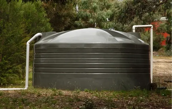 a large silver water collection storage tank with white pvc pipes connected sits on a dirt area with sparse green grass and green trees and red flowers in the background