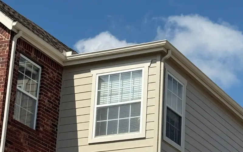 800x450 Corner of a residential apartment complex showing beige siding and red brick exterior walls with white-framed windows and white gutters along the roofline under a partly cloudy blue sky